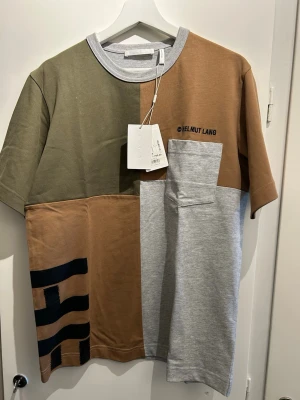 Patchwork t-shirt från Helmut Lang - Unik patchwork t-shirt från Helmut Lang i grått, brunt och olivgrönt. Stor svart logga baktill och mindre logga framtill. Kortärmad med rund hals och bröstficka i grått. Tillverkad i mjuk bomull för en chill och trendig look.