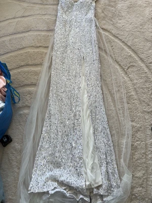 Vit glittrig långklänning med slits - Superelegant vit långklänning med glittriga paljetter och en hög slits framtill. Klänningen har en axelbandslös design och ett långt släp i tunn tyll som ger extra wow-känsla. Perfekt för dig som vill sticka ut på bal eller fest.