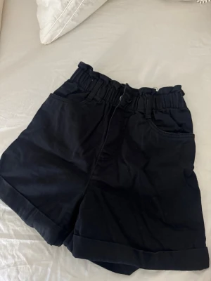 Svarta högmidjade shorts med resår - Snygga svarta shorts med hög midja och resår i midjan. Shortsen har uppvikta benslut, två framfickor och är tillverkade i ett mjukt bomullstyg. Perfekta för en avslappnad och trendig look.
