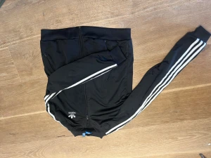 Svart Adidas ziptröja med vita ränder - Klassisk svart ziptröja från Adidas med vita ränder längs ärmarna och Adidas-logga på bröstet. Tröjan har dragkedja framtill, ståkrage och muddar vid ärmslut. Perfekt för en sportig och avslappnad stil.