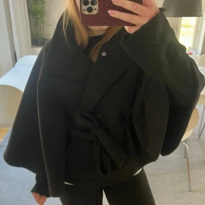 Svart cape-kappa med knytband - Stilren svart cape-kappa med bred krage och knytband i midjan. Har ändast provatttt