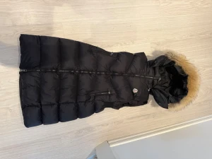 Svart lång dunväst med päls Moncler - Snygg svart lång dunväst från Moncler.. Västen har två dragkedjefickor, quiltad design och Moncler-logga på bröstet. Perfekt för lager på lager och håller dig varm under kyliga dagar.