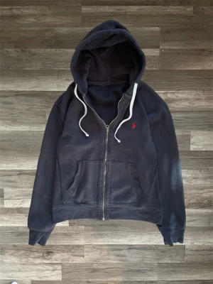 Mörkblå hoodie från Ralph Lauren - Säljer en mörkblå hoodie från Ralph Lauren med dragkedja och vit dragsko i huvan. Klassisk röd logga broderad på bröstet och två stora fickor framtill. Tillverkad i mjukt bomullsmaterial som är perfekt för chill dagar.