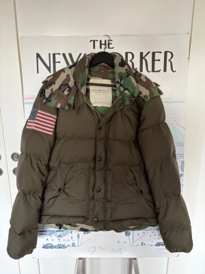 Grön dunjacka från RRL med camo-detaljer - Säljer en grön dunjacka från RRL med camouflage-detaljer på huvan och en stor amerikansk flagga på ärmen. Jackan har två framfickor med knappar och en hög krage. Perfekt för kalla vinterdagar och riktigt snygg streetstil.