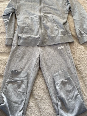 Grått loungewear-set från Puma - Mysigt grått loungewear-set från Puma med dragkedja på tröjan och två fickor framtill. Byxorna har resår i midjan och muddar vid bensluten. Setet är tillverkat i mjuk bomullsblandning och har Puma-logga på både tröja och byxa.