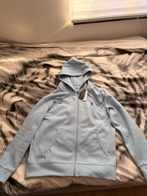 Polo kofta - Snygg ljusblå hoodie från Polo Ralph Lauren med dragkedja framtill och två fickor. Klassisk huva med dragsko och den ikoniska lilla loggan broderad på bröstet. Tillverkad i mjuk bomullsblandning som känns skön mot huden. Perfekt för en avslappnad och stilren look.