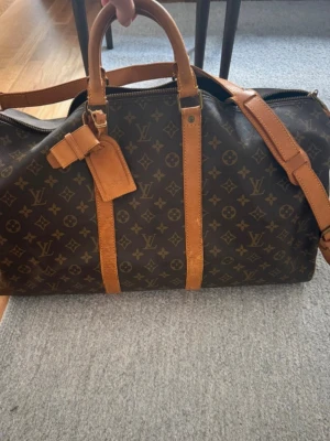 Louis Vuitton Monogram weekendväska - Säljer en stor och lyxig weekendväska från Louis Vuitton med klassiskt monogrammönster i brunt och beige. Väskan har läderdetaljer, dubbla handtag och en avtagbar axelrem. Perfekt för resor och har en tidlös design som sticker ut. Modell keepall 55