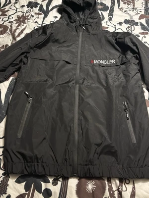 Svart Moncler vindjacka med huva - Svart vindjacka från Moncler med huva och dragkedja framtill. Jackan har två fickor med dragkedja och broderad Moncler-logga på bröstet samt ett broderat emblem på ärmen. Materialet är lätt och vattenavvisande, perfekt för blåsiga dagar.