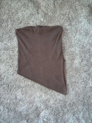 Brun ribbad asymmetrisk tubtopp - Säljer en brun ribbad tubtopp med asymmetrisk skärning. Toppen är ärmlös och har en cool, modern look som passar perfekt till jeans eller kjol. Materialet är stretchigt och bekvämt, vilket gör den enkel att styla för olika tillfällen.