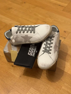Vita sneakers från Golden Goose - Stilrena vita sneakers från Golden Goose med grå stjärndetalj på sidan, grå skosnören och blå detalj vid hälen. Skorna har en klassisk låg siluett och är tillverkade i läder med gummisula. Perfekta för en trendig och avslappnad look.