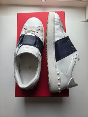 Valentino vita sneakers med svart detalj - Snygga vita sneakers från Valentino med bred svart läderdetalj över mitten och coola nitar på hälen. Skorna har en chunky sula och klassisk låg profil. Perfekta för dig som vill ha en stilren men ändå unik look. 100% äkta, allt og medföljer. Riktigt fint skick, endast något creasade och skav i hälen som inte syns eller känns vid användning.