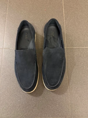Marinblå mocka loafers från ZARA - Snygga marinblå loafers från ZARA i mjuk mocka med stilren design. Skorna har en klassisk slip-on modell och vit sula som ger en fräsch kontrast. Perfekta för dig som gillar en clean och modern look till vardagsoutfiten.