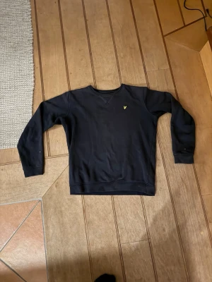 Tröja från Lyle & Scott - Snygg mörkblå sweatshirt från Lyle & Scott Junior med klassisk rund hals och gul logga på bröstet. Tröjan har ribbade muddar vid ärmslut och nederkant, perfekt för en avslappnad och stilren look.