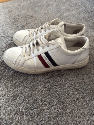  Moncler sneakers  - Snygga vita sneakers från Moncler med klassiska röda och marinblå ränder på sidan samt Moncler-logga. Skorna har lågt skaft, vita snören och är tillverkade i läder. Perfekta för dig som gillar stilrena och exklusiva sneakers.