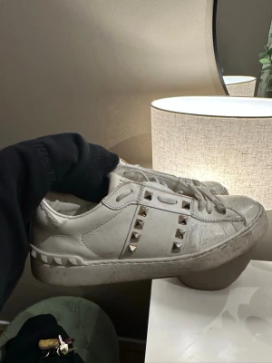 Vita sneakers med nitar från Valentino - Snygga vita sneakers från Valentino med coola silverfärgade nitar på sidorna. Skorna har en chunky sula och är tillverkade i läder. Klassisk låg modell med snörning och diskreta detaljer på hälen.
