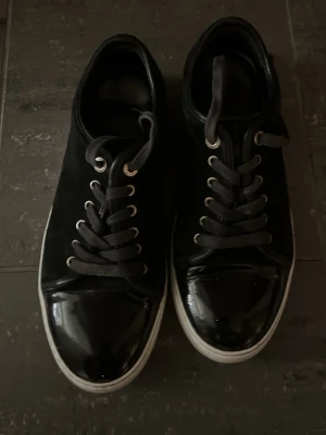 Svarta sneakers med lackdetaljer - Snygga svarta sneakers med glansiga lackdetaljer på tån och klassisk vit sula. Skorna har svarta skosnören och metallöljetter. Ovandelen är i mockaliknande material och tån i blankt material för en cool kontrast.