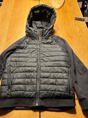 Svart pufferjacka från Superdry - Snygg svart pufferjacka från Superdry med quiltad framsida och baksida, mjuka svarta ärmar och huva. Jackan har dragkedja framtill, två sidofickor med dragkedja och ribbade muddar. Perfekt för kalla vinterdagar och riktigt skön att bära.