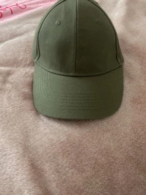 Olivgrön klassisk keps - Säljer en stilren olivgrön keps i bomull med böjd skärm och justerbart spänne bak. Kepsen har en enkel design utan logga eller tryck, perfekt för en clean och avslappnad look.