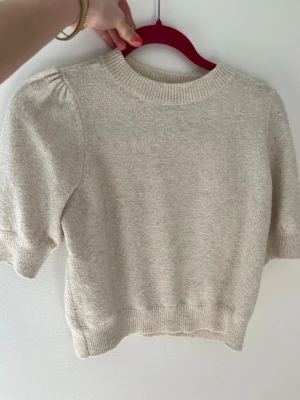 Beige stickad topp med puffärm - Snygg beige stickad topp med puffärmar och ribbade muddar vid hals, ärmslut och nederkant. Kortare modell med rund halsringning och mjukt material, perfekt för en trendig och avslappnad look.