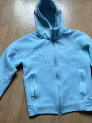 Nike X Nocta - Säljer en ljusblå hoodie från Nike x nocta med hel dragkedja framtill och två fickor med dragkedja. Hoodien har en klassisk huva och den ikoniska Nike-loggan broderad på bröstet. Perfekt för en sportig och avslappnad stil. Har använt den endast två gånger