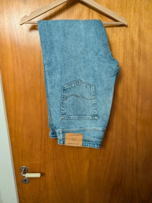 blå raka jeans Jack and Jones - Jeans i ljusblå denim med raka ben och femficksmodell. Jeansen har knappgylf och en snygg vintage-look. Perfekta för dig som gillar tidlös stil och vill ha ett par ikoniska jeans i garderoben.