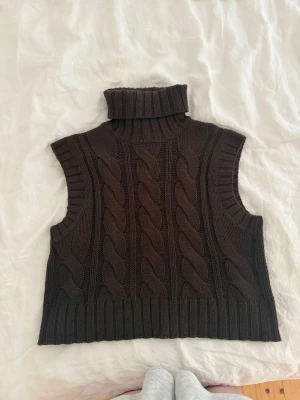 Mörkbrun stickad väst med polokrage - Snygg mörkbrun stickad väst med polokrage och grovt flätmönster framtill. Ärmlös modell med ribbade kanter och mjukt material, perfekt att styla över en skjorta eller långärmad tröja.