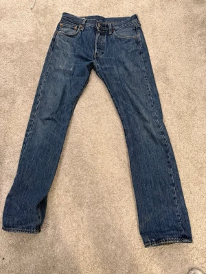 Klassiska blå Levi's jeans straight fit - Säljer ett par klassiska blå Levi's jeans med rak passform och fem fickor. Jeansen har en mörkblå tvätt, orange sömmar och läderpatch bak i midjan. Tillverkade i slitstark denim, perfekt för en avslappnad och tidlös stil.
