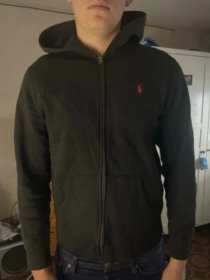 Svart hoodie från Ralph Lauren - Säljer en svart hoodie med dragkedja från Ralph Lauren. Klassisk design med huva, fickor framtill och den ikoniska röda loggan broderad på bröstet. Perfekt för dig som gillar stilrena och bekväma plagg.