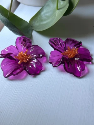 Stora lila blomörhängen med pärlor - Unika örhängen formade som stora blommor i transparent lila plast med orange pärlor i mitten. Perfekta för dig som vill sticka ut och addera färg till din stil. Blommornas detaljer ger ett coolt och lekfullt intryck.