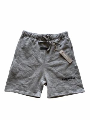Grå shorts från Essentials - Gråa shorts från Essentials med snörning i midjan och tryckt logga på vänster ben. Mjukt material och avslappnad passform, perfekta för chill dagar eller när du vill vara bekväm. Klassisk design med sidofickor.