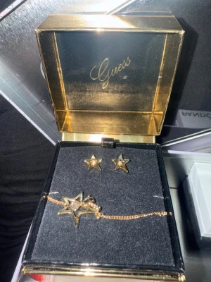 Guess smyckesset med stjärnor - Snyggt smyckesset från Guess med örhängen och armband i guldton. Båda delarna har stjärnformade hängen med glittrande stenar. Perfekt för dig som gillar trendiga accessoarer med lite extra bling.