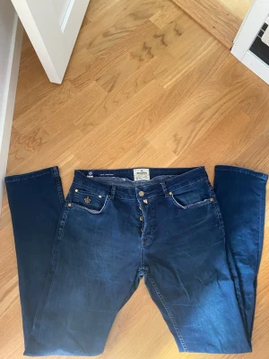 Mörkblå Steve Satin Jeans 34/34 - Mörkblå jeans från Steve Satin Jeans med klassisk femficksdesign och guldfärgade nitar. Snygg detalj med broderad lilja på myntfickan. Raka ben och normal midja. Perfekta för en trendig och avslappnad look.