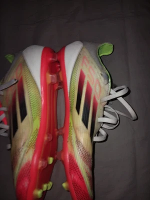 Adidas F50 färgglada fotbollsskor - Säljer ett par Adidas F50 fotbollsskor med snygg design i vitt, neonlime, svart och rött. Skorna har klassiska svarta Adidas-ränder på sidan, snörning och dobbar för bra grepp på planen. Ovandelen är i syntetmaterial och insidan har neonfärgade detaljer.