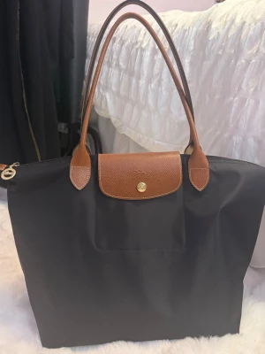 Svart Longchamp Le Pliage handväska Large - Klassisk svart Le Pliage handväska från Longchamp med bruna läderdetaljer och handtag. Skickar även en innerväska. Använd bara 2-3 ggr.