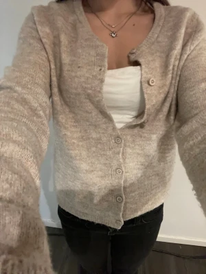 Beige stickad kofta med knappar - Mysig beige stickad kofta med långa ärmar och rund halsringning. Koftan har flera knappar framtill och en avslappnad passform. Perfekt att slänga över en topp för en chill look.