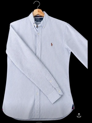 Randig skjorta från Polo Ralph Lauren - Klassisk slim fit skjorta från Polo Ralph Lauren i ljusblått och vitt med tunna ränder. Skjortan har button-down krage, långa ärmar och broderad logga på bröstet. Tillverkad i bomull och stängs med knappar framtill.