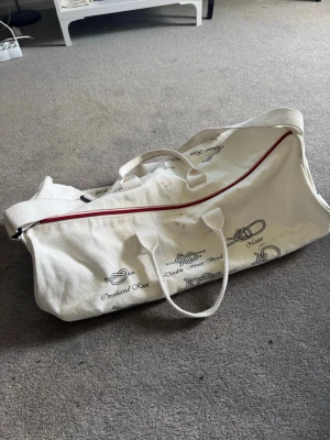 Vit treven bag från sebago  - Stor vit shoppingväska i canvas med tryck av olika knutar och svart text. Väskan har två handtag, en axelrem och röd dragkedja upptill. Perfekt för att bära med sig mycket grejer och sticker ut med sitt unika mönster.
