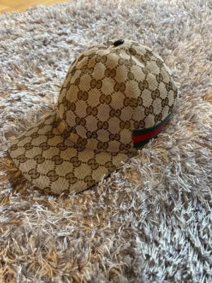 Gucci keps med GG-mönster - Snygg keps från Gucci i beige canvas med klassiskt brunt GG-monogram över hela kepsen. Grön och röd rand på sidan och justerbart spänne bak i läder. Perfekt accessoar för en trendig streetstyle-look. Självklart äkta !