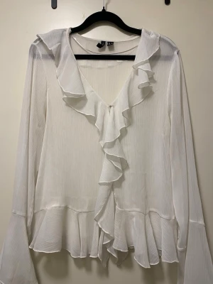 Vit volangblus från VeroModa - Stilren vit blus från VeroModa med långa ärmar och volangdetaljer längs urringningen och framtill. Blusen har en lätt transparent känsla och ett mjukt, flytande tyg som ger en romantisk look. Perfekt att styla med jeans eller kjol. Storlek M men passar S perfekt 
