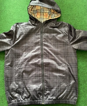 Svart vindjacka med rutigt foder - Stilren svart vindjacka från Burberry med diskret rutmönster på utsidan och klassiskt beige, röd och blått rutigt foder. Jackan har huva, dragkedja framtill och två sidofickor. Perfekt för blåsiga dagar och enkel att matcha med olika outfits.