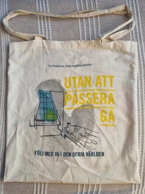 Tygkasse Kriminalvården - Beige tygkasse i bomull med tryck från Kriminalvårdens poddserie 'Utan att passera gå'. Ena sidan har illustration och text i gult och blått, andra sidan har citat och grafiskt motiv. Långa handtag, enkel och rymlig modell. Någon enstaka lite fläck.