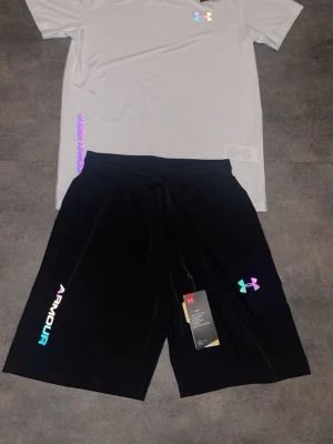 Under Armour set med t-shirt och shorts - Snyggt träningsset från Under Armour med en ljusgrå t-shirt och svarta shorts. Båda plaggen har holografiska loggor och textdetaljer. Tillverkade i ett lätt och snabbtorkande material som passar perfekt till träning eller sport.