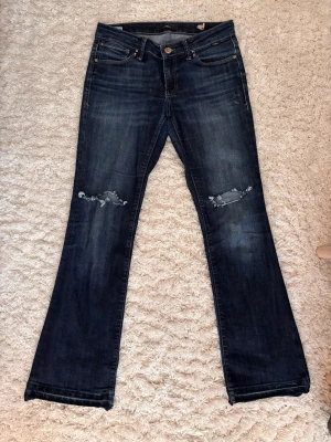 Lågmidjade jeans - Lågmidjade stretchiga lösa bootcut jeans från Mavi köpta på Zalando för 900kr. Klippt egna hål över knäna. Storlek 29/30 vilket motsvarar en S ungefär och dem passar någon som är 160-165cm. Bottensömmen är uppsprättad för att bli längre. 