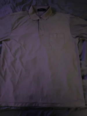 Beige pikétröja med bröstficka från Hugo boss - Klassisk beige pikétröja från Hugo boss med krage och knappar framtill. Tröjan har en bröstficka på vänster sida och är tillverkad i ett mjukt bomullstyg. Perfekt för en avslappnad och stilren look.