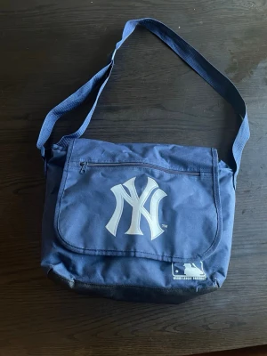 NY Yankees “book bag” - Snygg mörk blå axelväska i canvas med stort New York Yankees-tryck på locket och justerbar axelrem. Väskan har dragkedjeficka framtill och MLB-logga nedtill. Perfekt för dig som gillar laid back cool stil eller bara snygga väskor! Ställ frågor om ni vill! (pssst pris kan diskuteras)