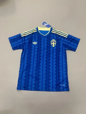 Sveriges Vm tröja blå - Blå fotbollströja från Adidas med Sveriges landslagsemblem på bröstet. Tröjan har gula detaljer, tre ränder på axlarna och ett mönster i ljusblått. Tillverkad i polyester och har korta ärmar samt rund hals. Storlek M