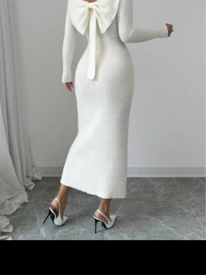 Vit stickad långklänning med rosett - Elegant vit stickad långklänning med lång ärm och stor dekorativ rosett i ryggen. Klänningen har en kroppsnära passform och är tillverkad i ett ribbat material som ger en snygg siluett.