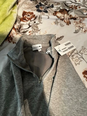 Grå halvzip hoodie från Lacoste - Snygg grå halvzip hoodie från Lacoste med klassisk krokodillogga på bröstet. Tröjan har hög krage, långa ärmar och ribbade muddar. Tillverkad i mjukt material som känns skönt mot huden. Perfekt för en avslappnad och stilren look.