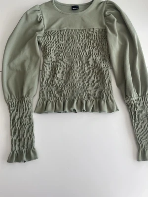 Grön smockad topp från Gina Tricot - Superfin ljusgrön topp från Gina Tricot med smockad kropp och ärmslut. Långa puffärmar och volangdetalj nertill ger en trendig look. Perfekt att styla med jeans eller kjol för en cool vibe.