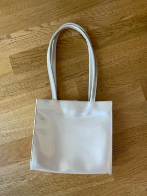 Glansig beige handväska från Escada - Stilren Glansig beige handväska från Escada i blankt material med fyrkantig form och dubbla axelband. Väskan har minimalistisk design med diskret logga framtill och passar perfekt för dig som gillar en clean look.